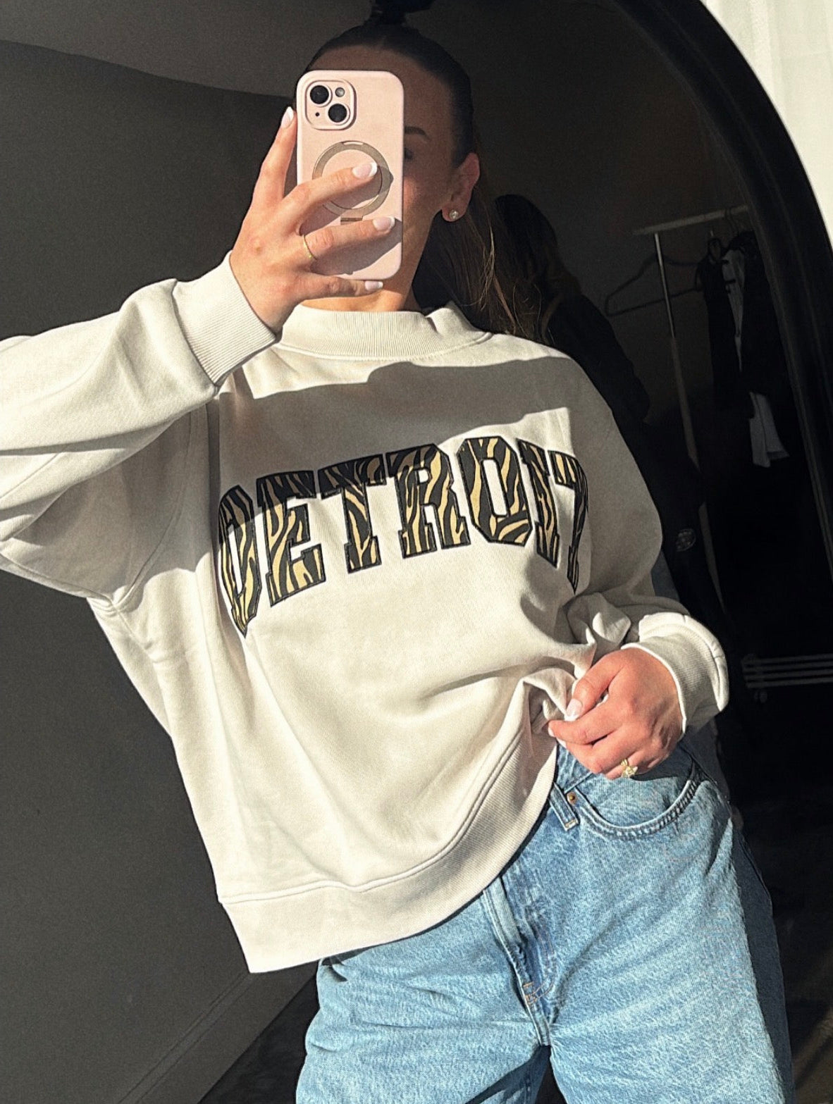 Person wearing a white sweatshirt with text and blue jeans, taking a mirror selfie.