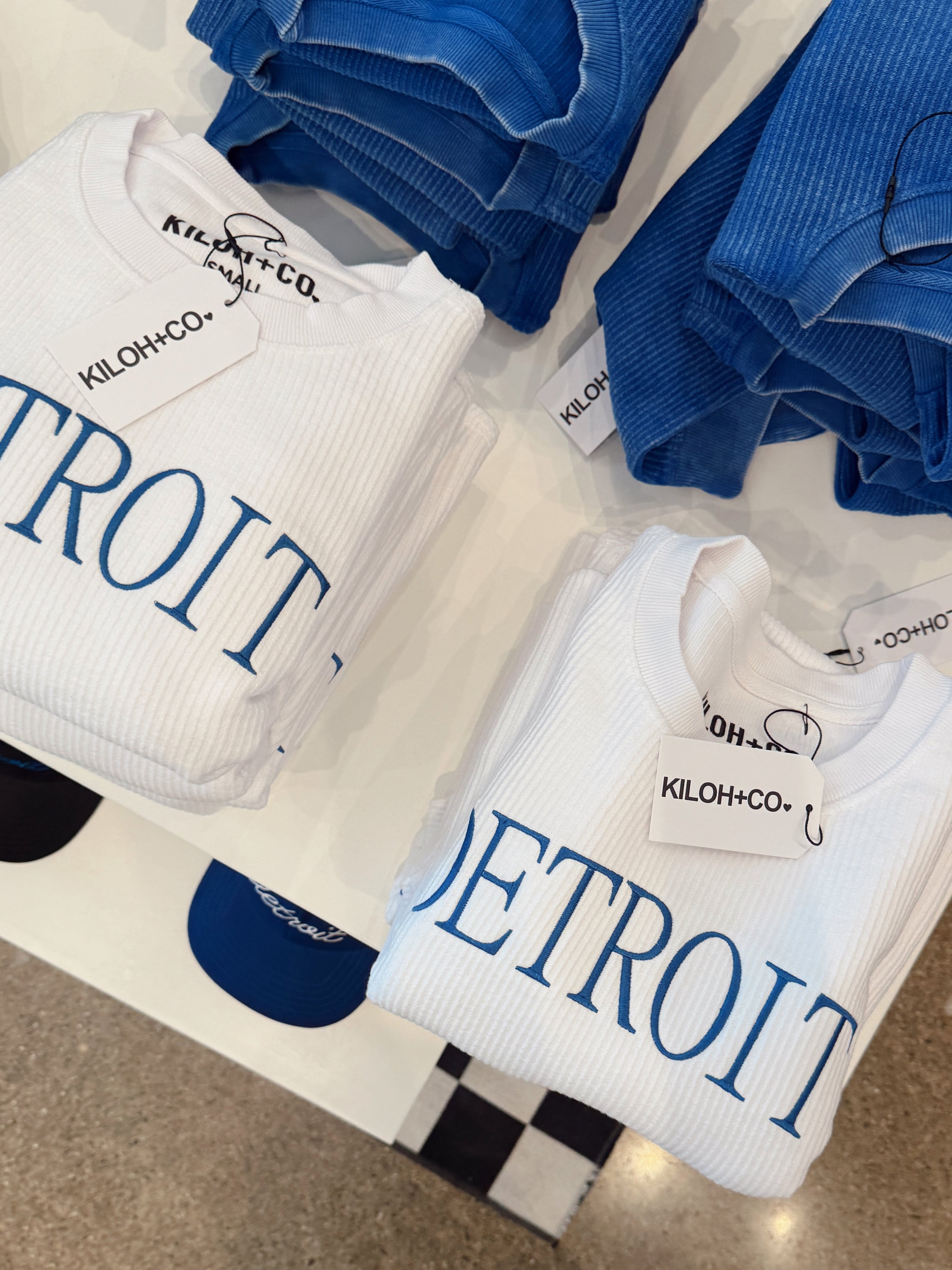 White sweatshirts with 'Detroit' printed on them, displayed with blue pants and accessories.